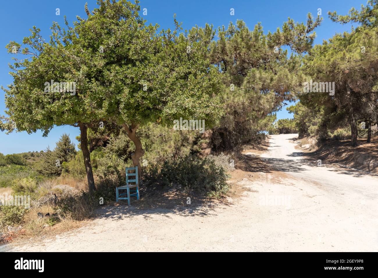 Landstraße in den Bergen von Kefalos mit einem blauen Stuhl im Schatten unter einem Baum. Abseits der ausgetretenen Pfade. Kos, Dodekanes, Griechenland Stockfoto
