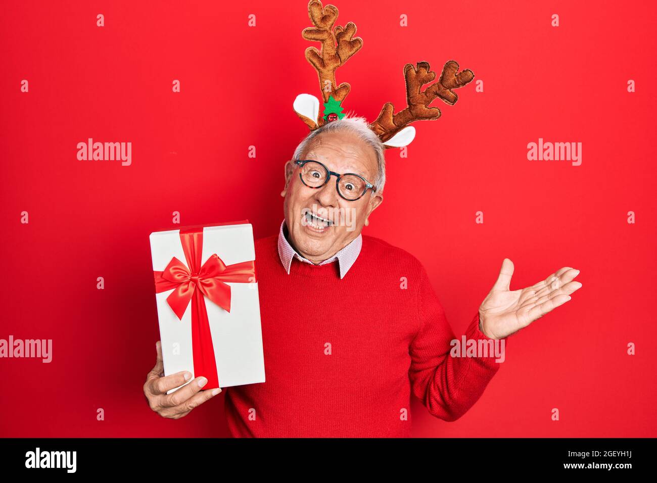 Senior Mann mit grauen Haaren trägt Hirsch weihnachtshut mit Geschenk feiern Leistung mit glücklichem Lächeln und Gewinner Ausdruck mit erhobener Hand Stockfoto