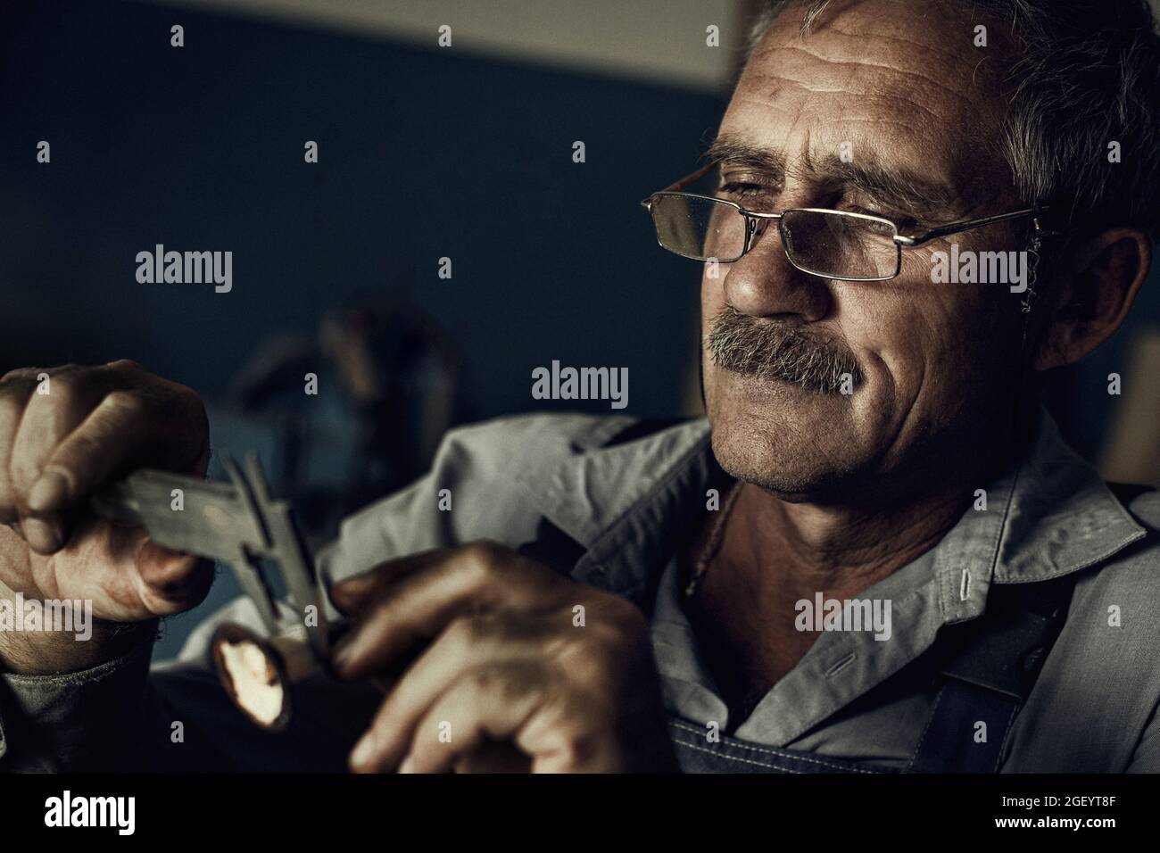 Ein alter Mann mit Brille bei der Arbeit in der Werkstatt misst ein Detail. Künstlerisches Foto eines älteren Meisters. Filmische Verarbeitung der Fotografie. Stockfoto