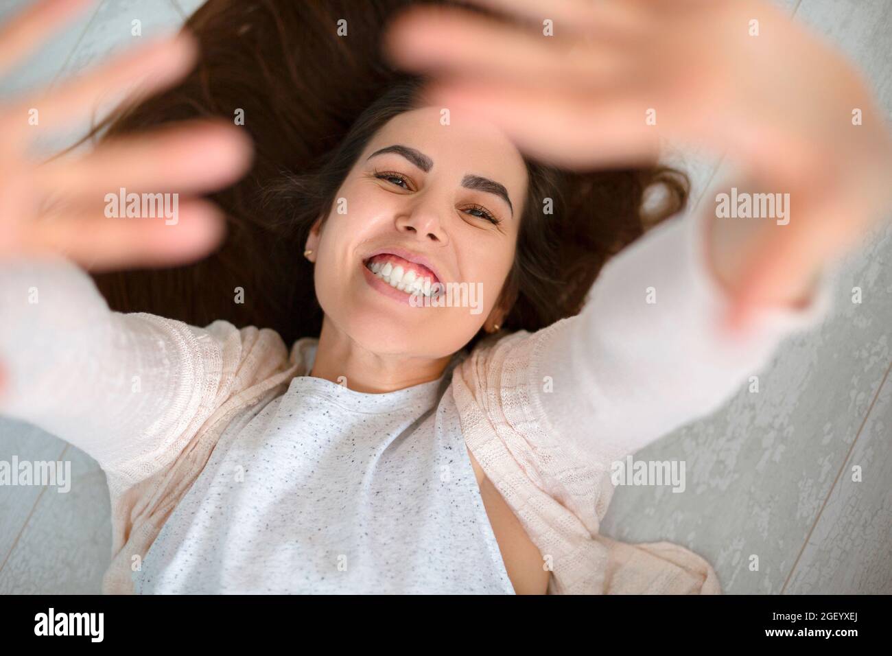 Porträt von oben einer glücklichen jungen positiven Frau, die auf einem rustikalen Holzboden liegt und beide Hände hochhebt und mit einem breiten strahlenden Lächeln in die Kamera blickt Stockfoto