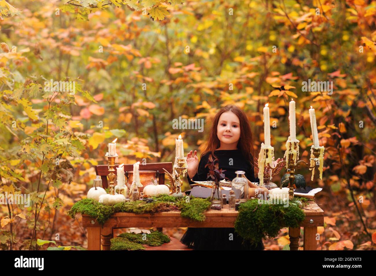 Kleines entzückendes Mädchen im schwarzen Hexenkostüm, das im Herbstwald spielt, Kind, das sich am Holztisch mit Moos, Kürbissen und verschiedenen Dekorationen amüsieren kann, lo Stockfoto