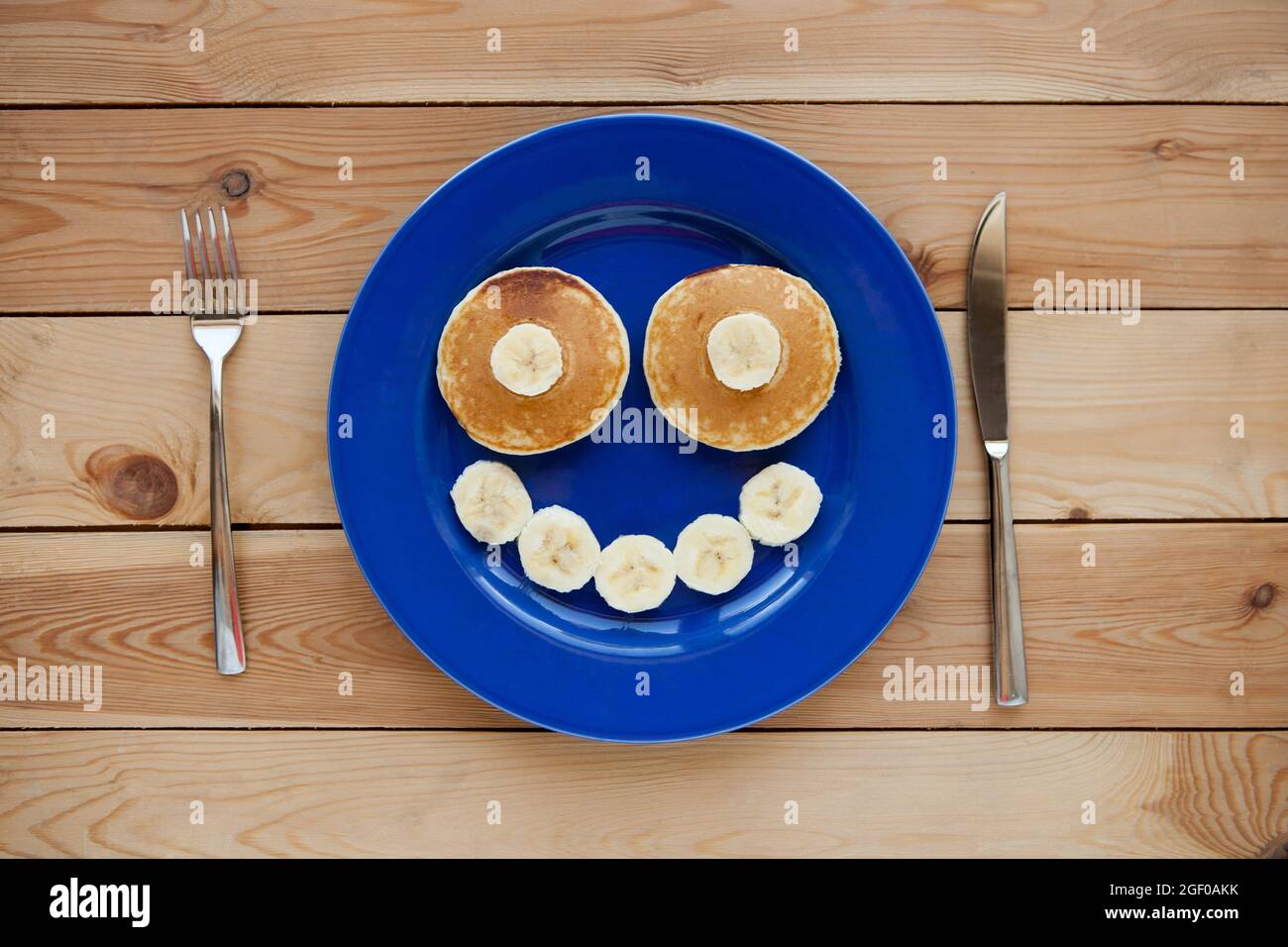 Pfannkuchen mit Lächeln auf blauem Teller und Holzhintergrund. Banana Fruit Smiling - lustiges Essen für Kinder Stockfoto