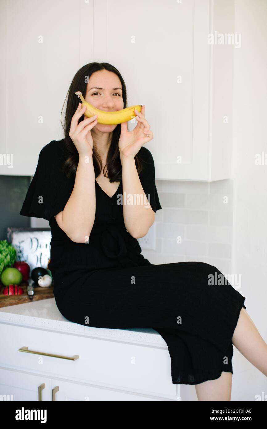 Eine kaukasische Frau mit einem schwarzen Kleid sitzt auf den Küchenmöbeln mit einer Banane auf dem Mund Stockfoto