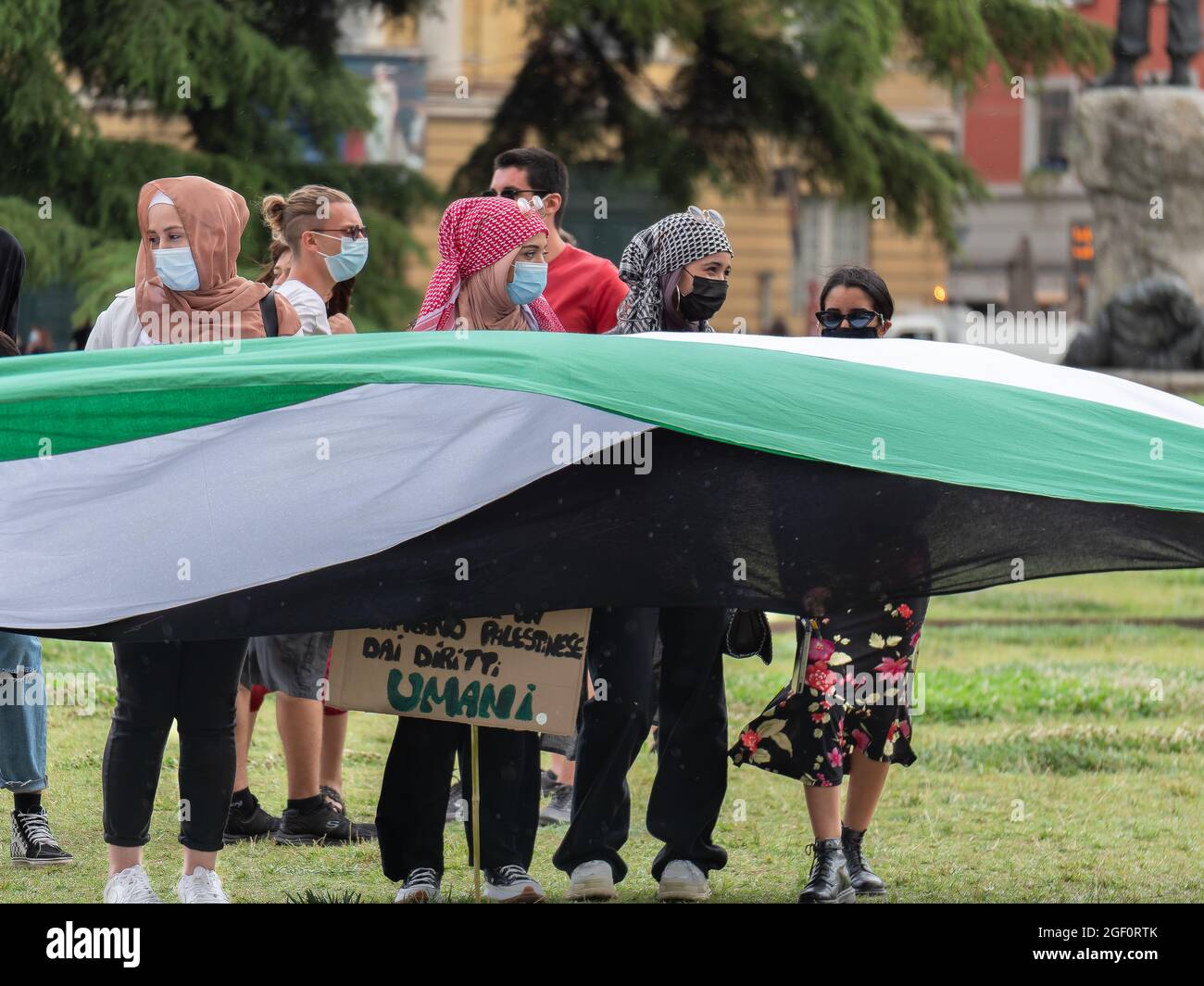 Arabische Mädchen mit chirurgischen Masken auf ihren Gesichtern stehen nebeneinander und schütteln eine große Flagge. Stockfoto