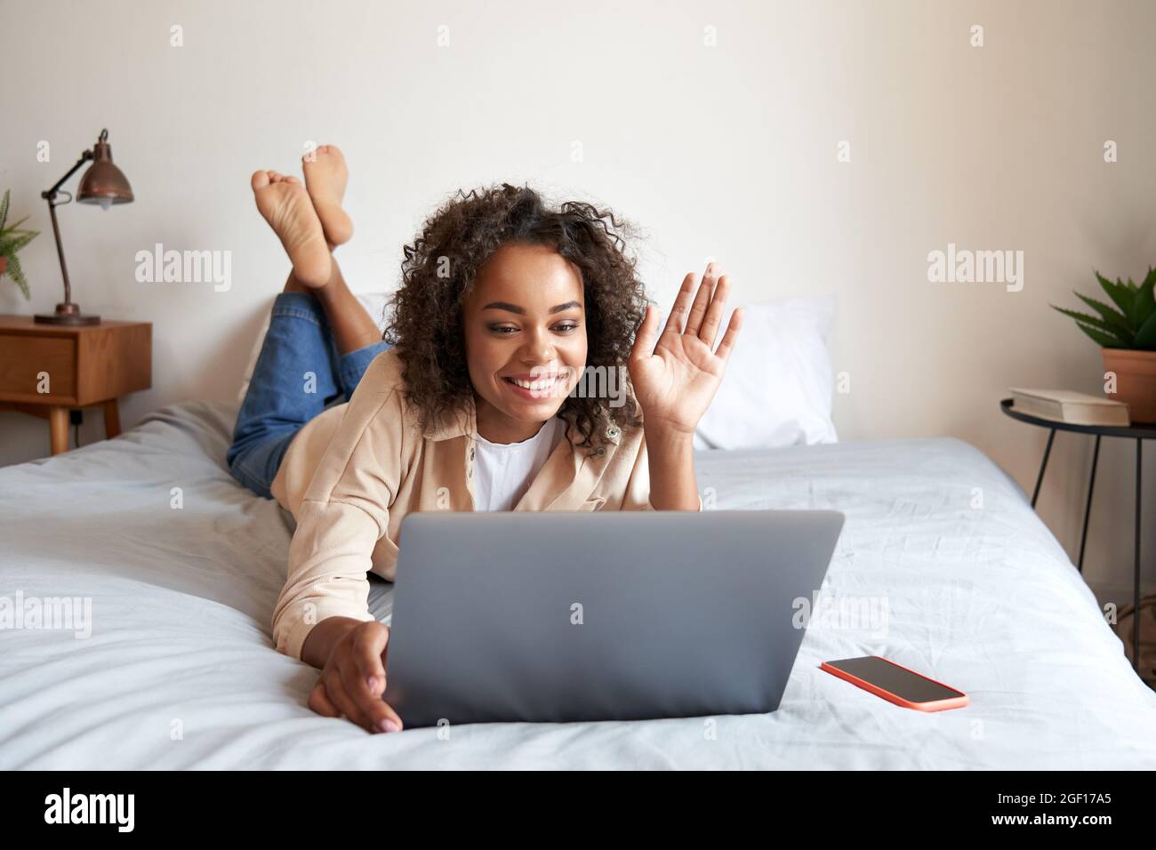 Junge afrikanische Frau, die auf dem Bett liegt, hat virtuelle Treffen, die mit dem Laptop die Hand winken. Stockfoto