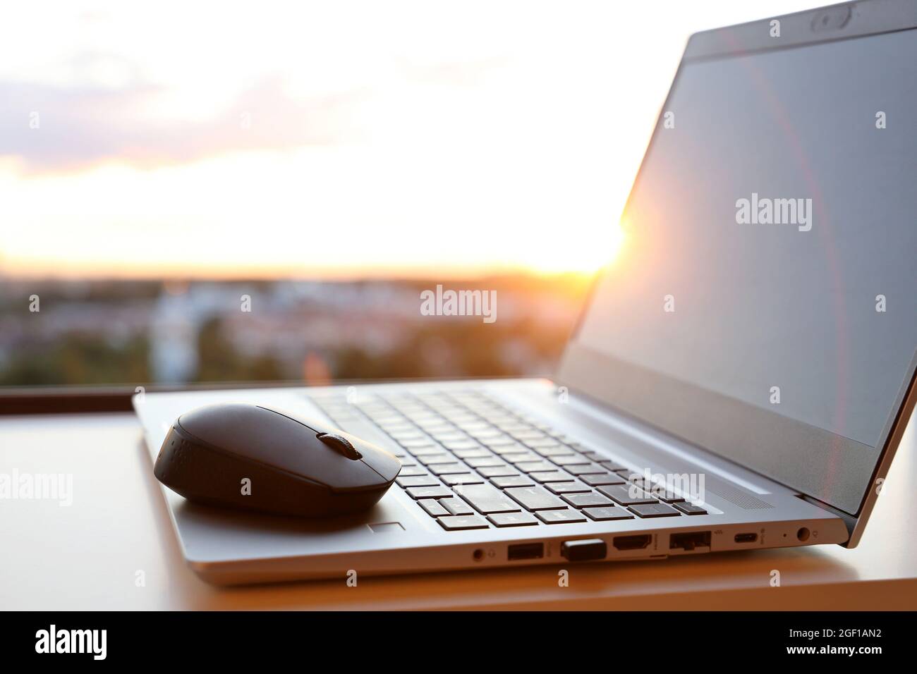 Laptop und kabellose Maus bei Sonnenaufgang auf dem Tisch gegen das Fenster. Gemütlicher Arbeitsplatz im Home Office, Konzept der Arbeit am frühen Morgen Stockfoto