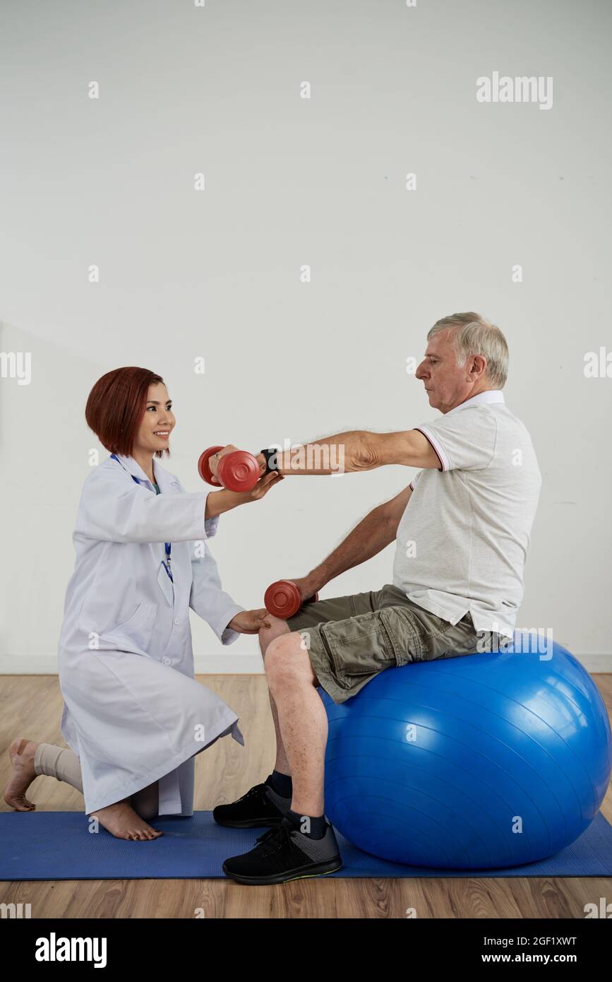 Der Physiotherapeut bittet den älteren Mann, der auf dem Fitnessball sitzt, die Hand mit einer Kurzhantel zu heben, um ein Rehabilitationskonzept zu entwickeln Stockfoto
