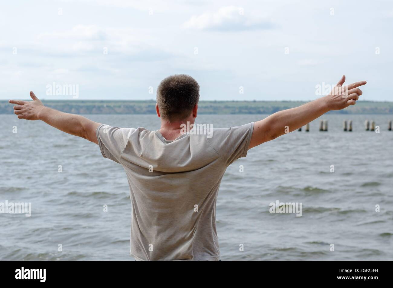 Halblanges Porträt eines erwachsenen Mannes, der am Ufer eines breiten Flusses steht. Mann in einem grauen T-Shirt mit kurzen Ärmeln, die Arme an den Seiten ausgestreckt. Re Stockfoto