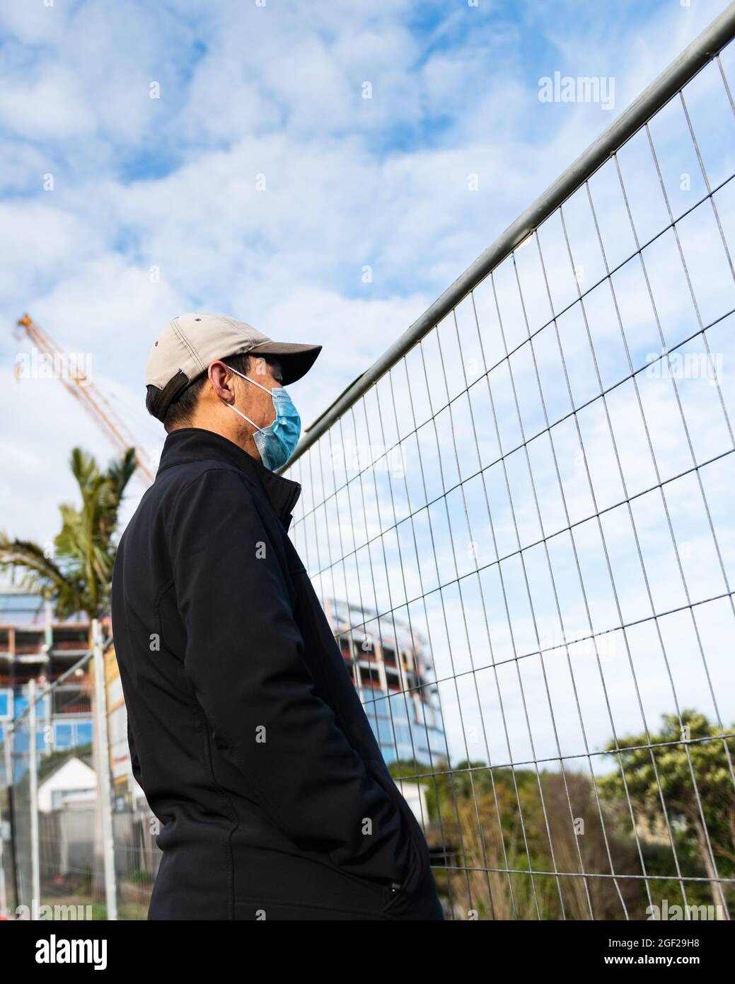 Mann mit Gesichtsmast, der durch die verdrahhte Industriezaunwand auf die Baustelle schaut. Vertikales Format. Stockfoto