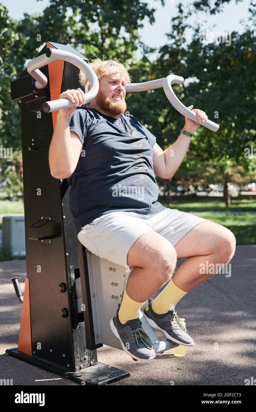 Übergewichtiger Mann, der auf Sportgeräten trainiert, hebt Gewichte und baut seine Muskeln im Freien auf Stockfoto