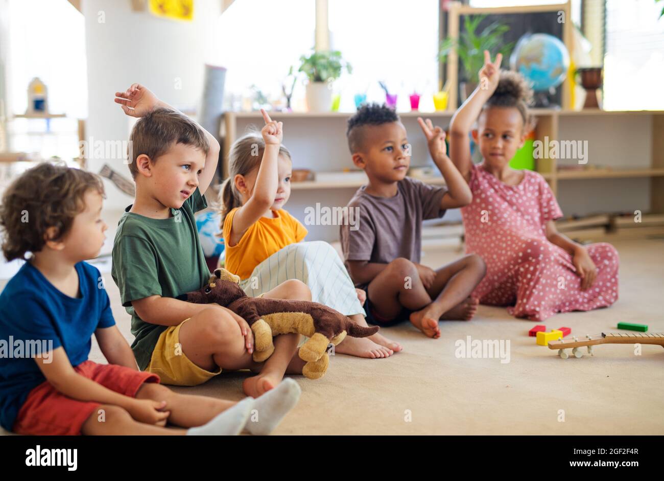 Gruppe von kleinen Kindergartenkindern, die im Klassenzimmer auf dem Boden sitzen und die Hände heben. Stockfoto