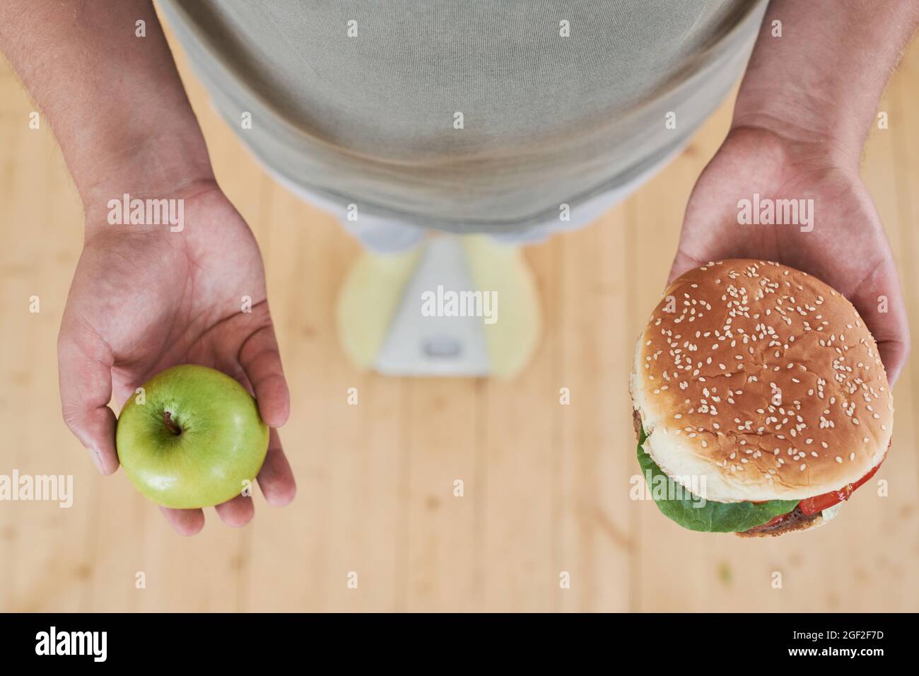 Nahaufnahme eines Mannes, der auf einer Waage steht und apfel in der einen Hand und Burger in der anderen Hand hält, um eine schwierige Wahl zu treffen Stockfoto
