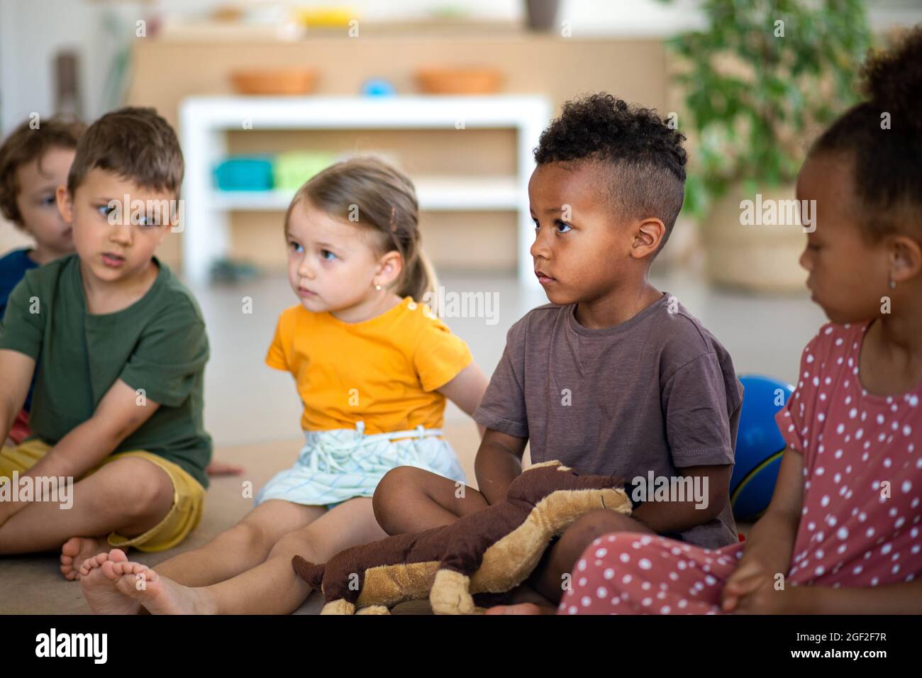 Gruppe von kleinen Kindergartenkindern, die im Klassenzimmer auf dem Boden sitzen und dem Lehrer zuhören. Stockfoto