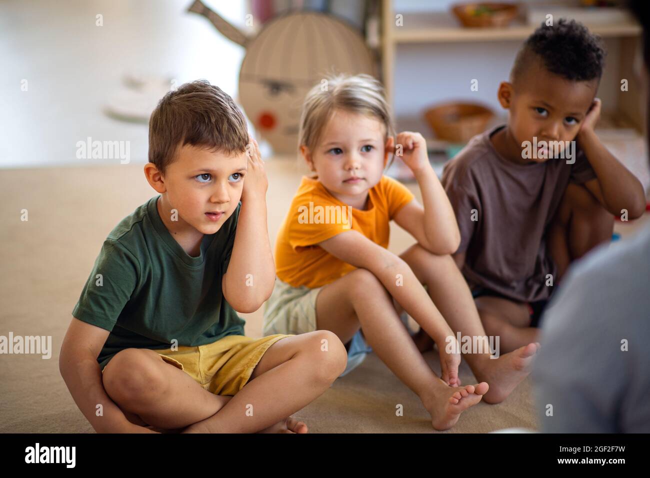 Gruppe von kleinen Kindergartenkindern, die im Klassenzimmer auf dem Boden sitzen und dem Lehrer zuhören. Stockfoto