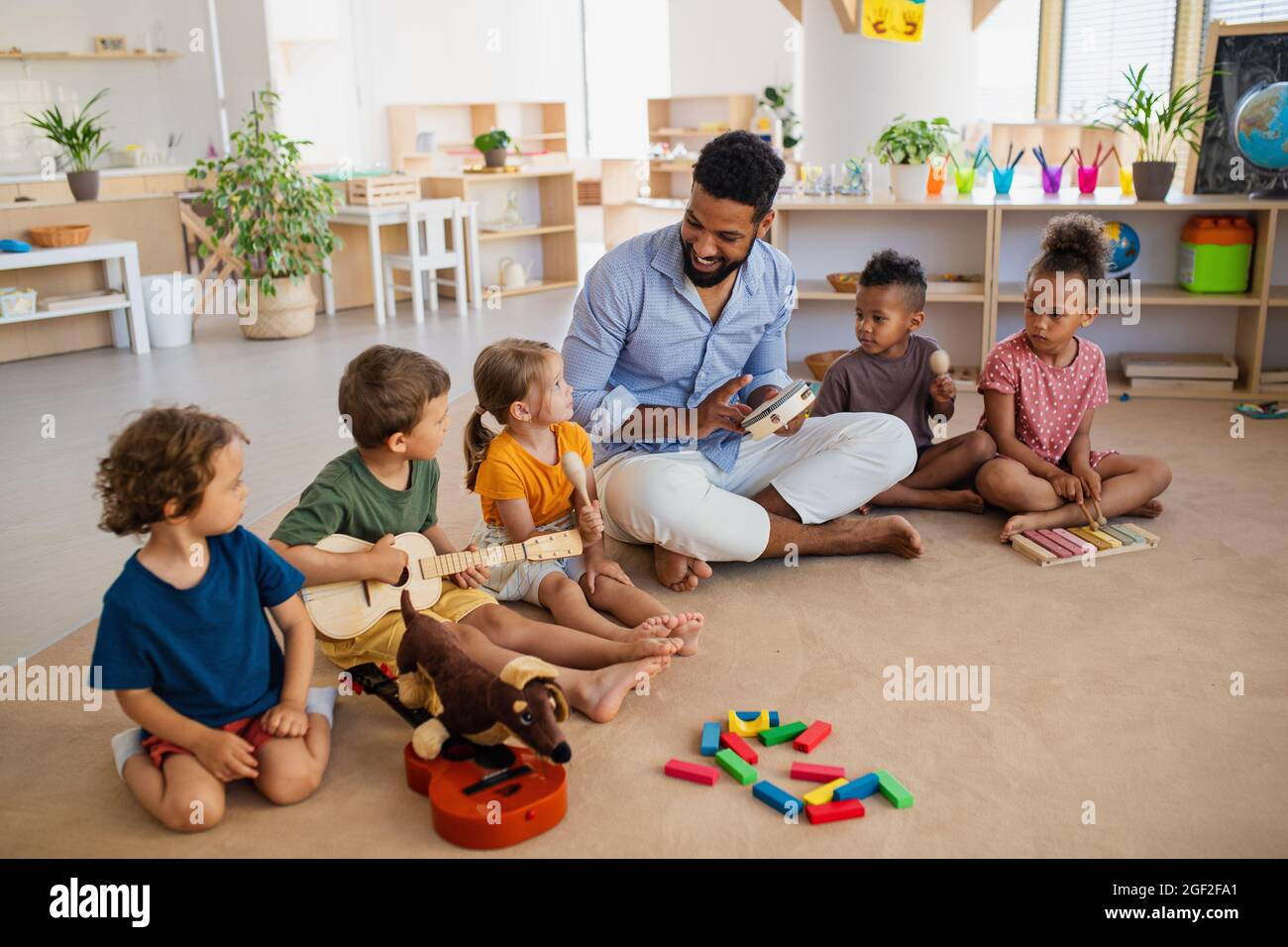 Gruppe von kleinen Kindern im Kindergarten mit einem Lehrer, der im Klassenzimmer auf dem Boden sitzt und Musikinstrumente spielt. Stockfoto