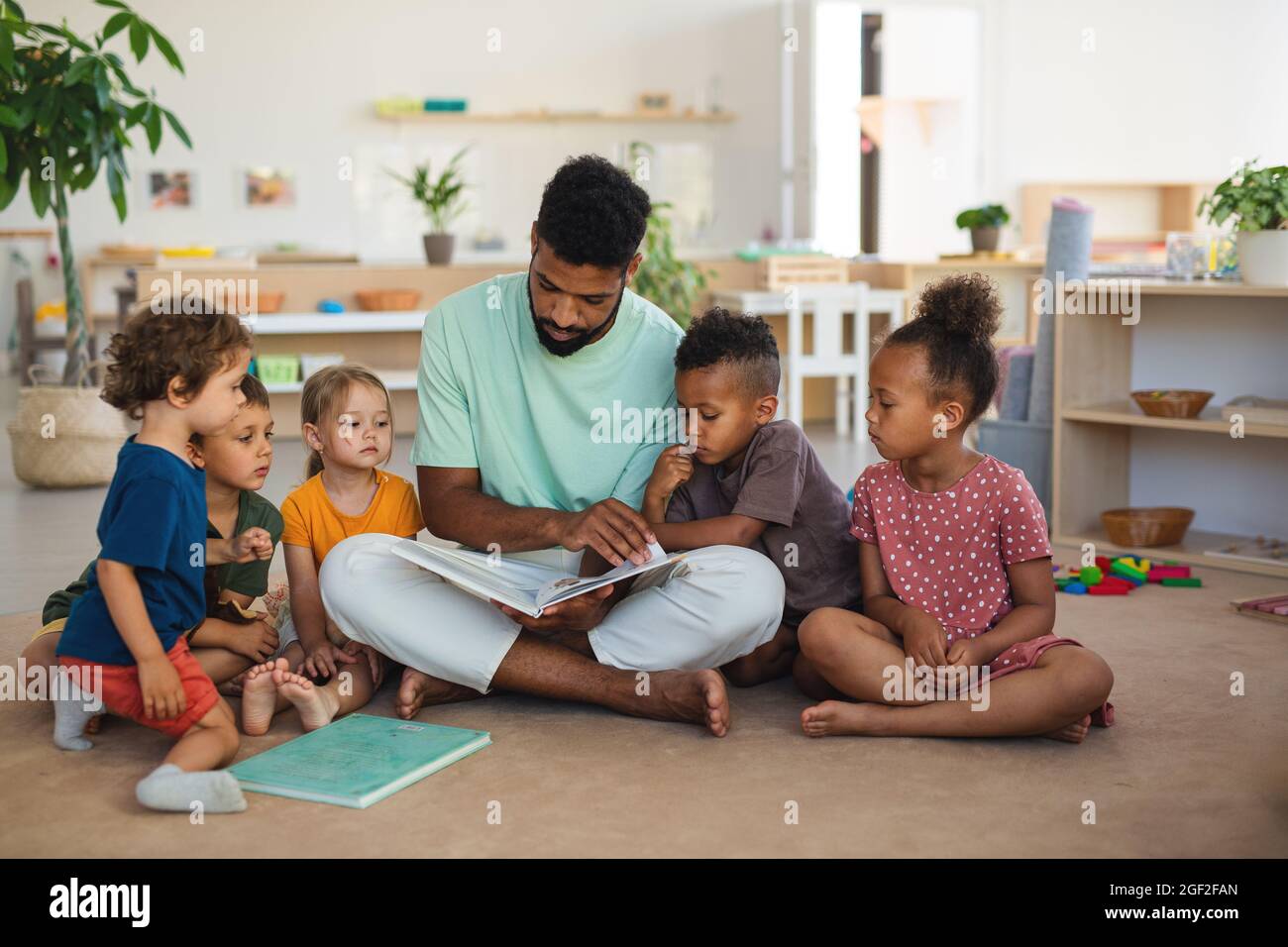 Gruppe von kleinen Kindergartenkindern mit einem Lehrer, der drinnen im Klassenzimmer auf dem Boden sitzt und ein Märchenbuch liest. Stockfoto