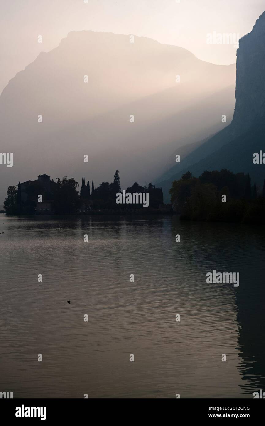 Sonnenuntergang im September über einem alpinen See in Norditalien, dem Lago di Toblino in Trentino-Südtirol. Die mittelalterliche Burg Castel Toblino ist in Silhouette gehalten, da die Balken der sinkenden Sonne die zerklüftete Landschaft zwischen den Dolomiten von Monte Casale und Monte Garzole oder Piccolo Dain beleuchten. Stockfoto