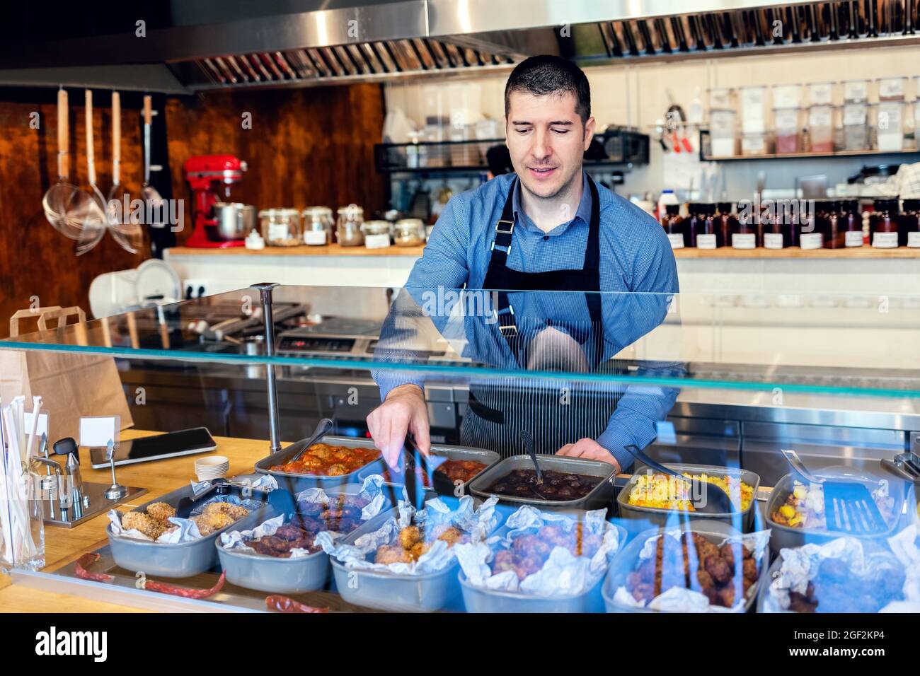 Ein zufriedener Kellner mit Schürze servierte den Gästen Essen zum Mitnehmen an der Theke in einem kleinen Familienrestaurant. Stockfoto