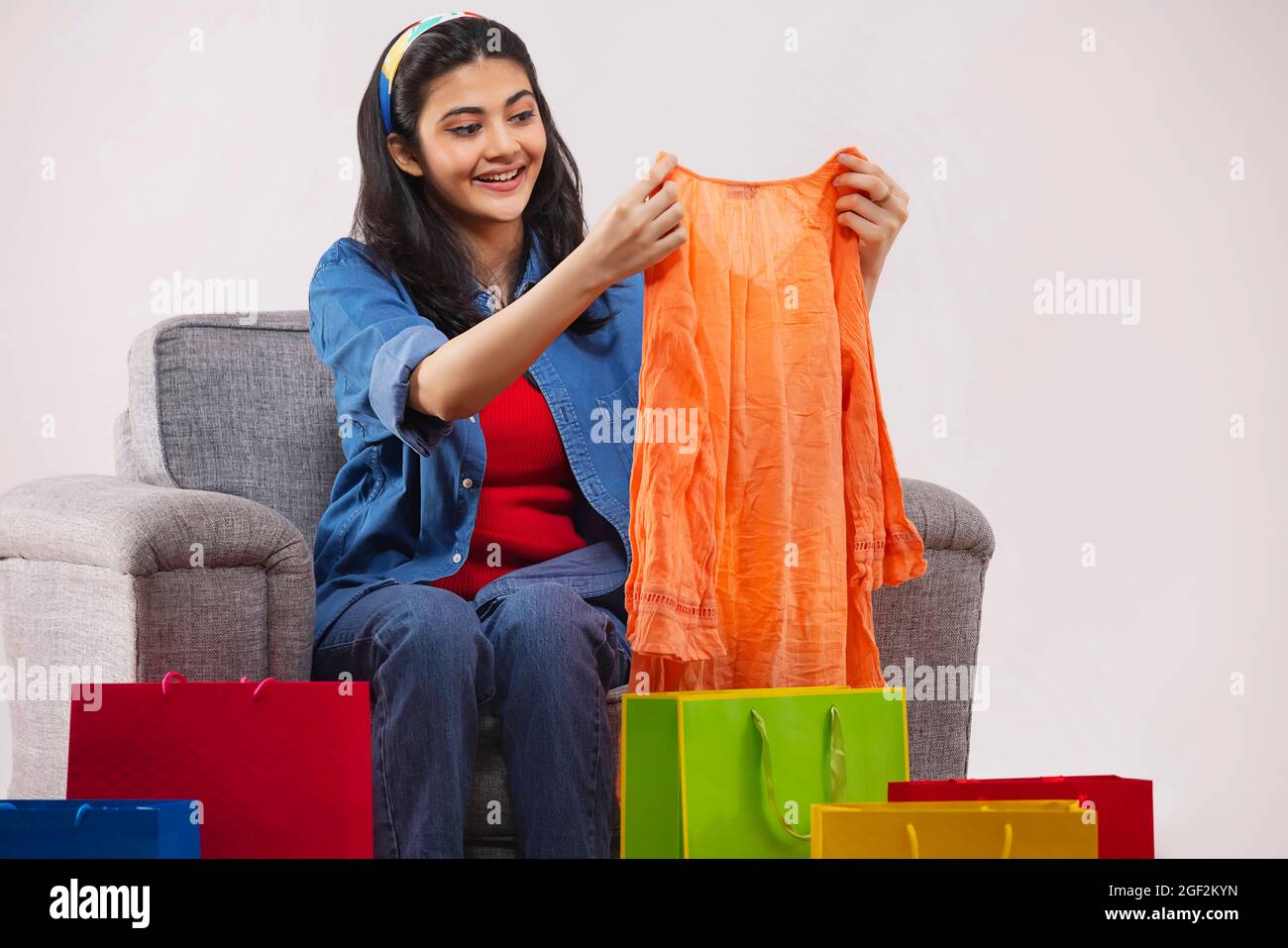 Eine junge Frau, die ihr neues Kleid auspackend, sitzt auf dem Sofa inmitten farbenfroher Einkaufstaschen in ihrem Zimmer. Stockfoto