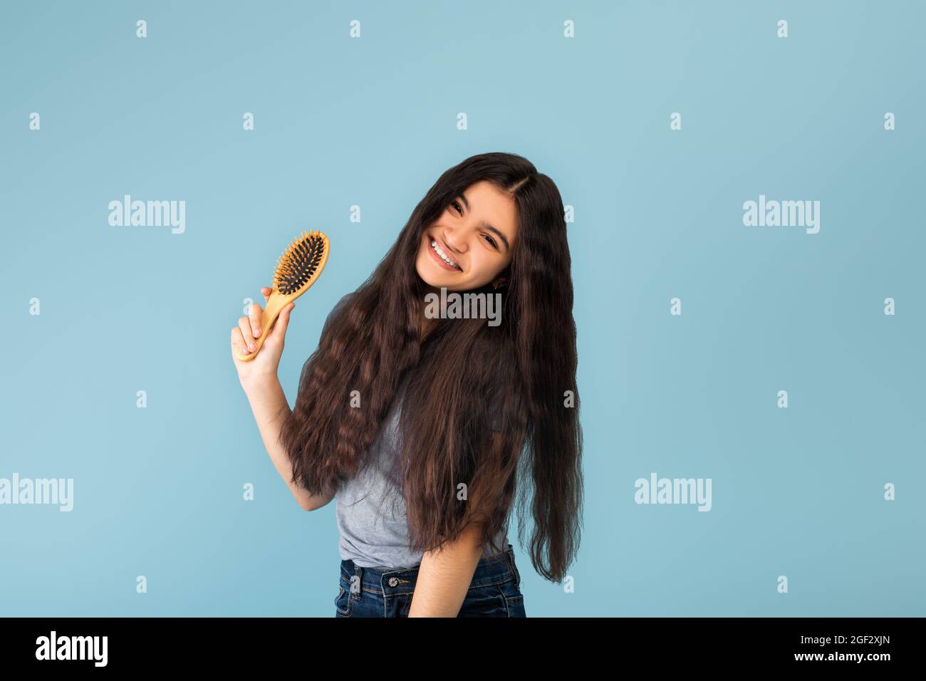 Attraktive indische Teenager-Mädchen mit langen seidig dunklen Haaren halten Holzbürste, posiert über blauen Studio-Hintergrund Stockfoto