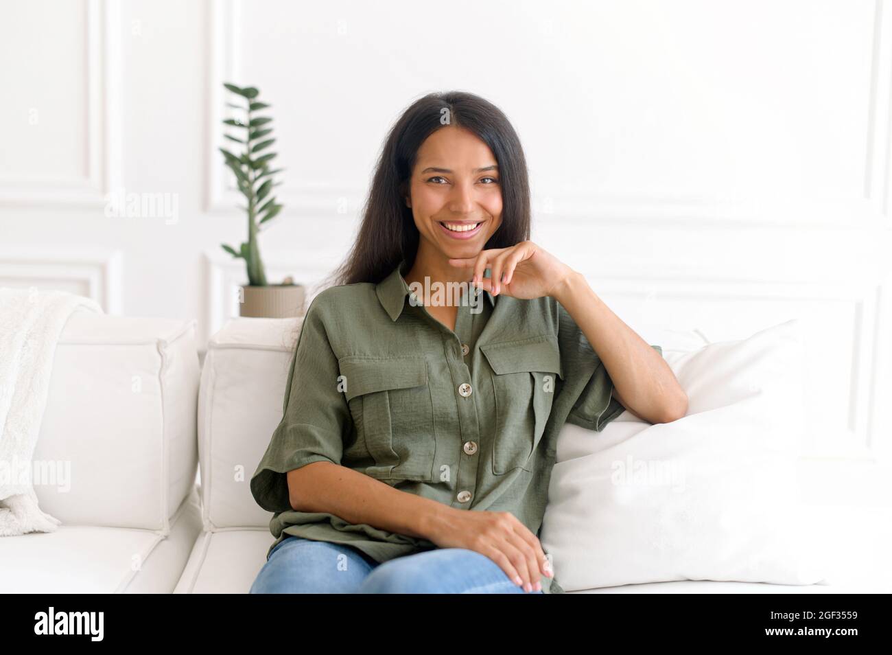 Porträt einer ruhigen und charmanten schönen indischen dunkelhaarigen Frau. Die hübsche Mixed-Race-Dame in legerer Kleidung blickt auf die Kamera und lächelt, auf dem bequemen weißen Sofa sitzend Stockfoto