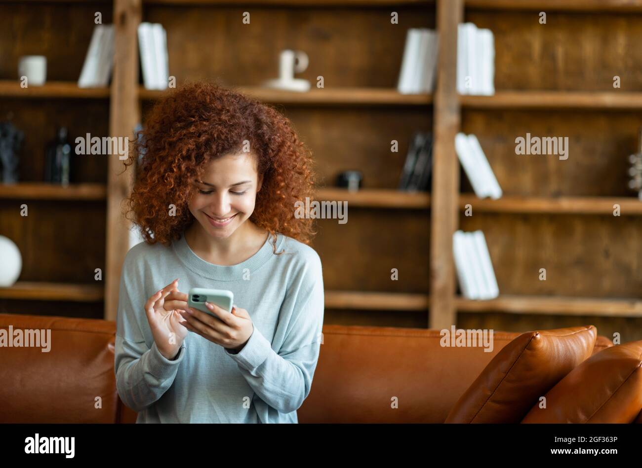 Fröhliche junge rothaarige lockige Frau chattet gerne online, sitzt zu Hause auf der Couch und Messaging auf dem Smartphone. Lächelnde Frau verbringt Freizeit in sozialen Netzwerken, scrollende Feed Stockfoto