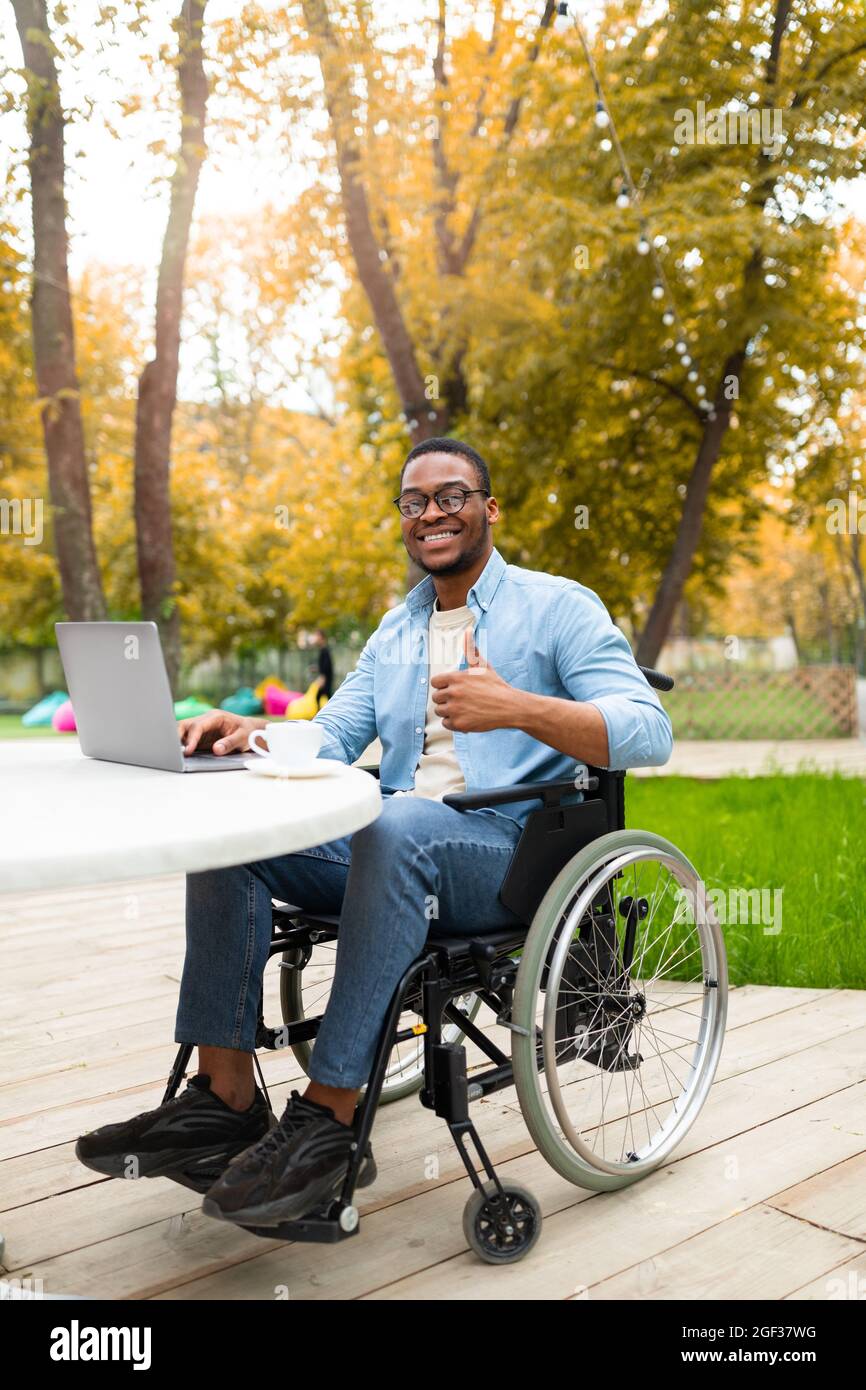 Online-Arbeit für behinderte Menschen. Schwarzer Mann im Rollstuhl, der am Laptop arbeitet und im Café im Herbstpark Daumen zeigt Stockfoto