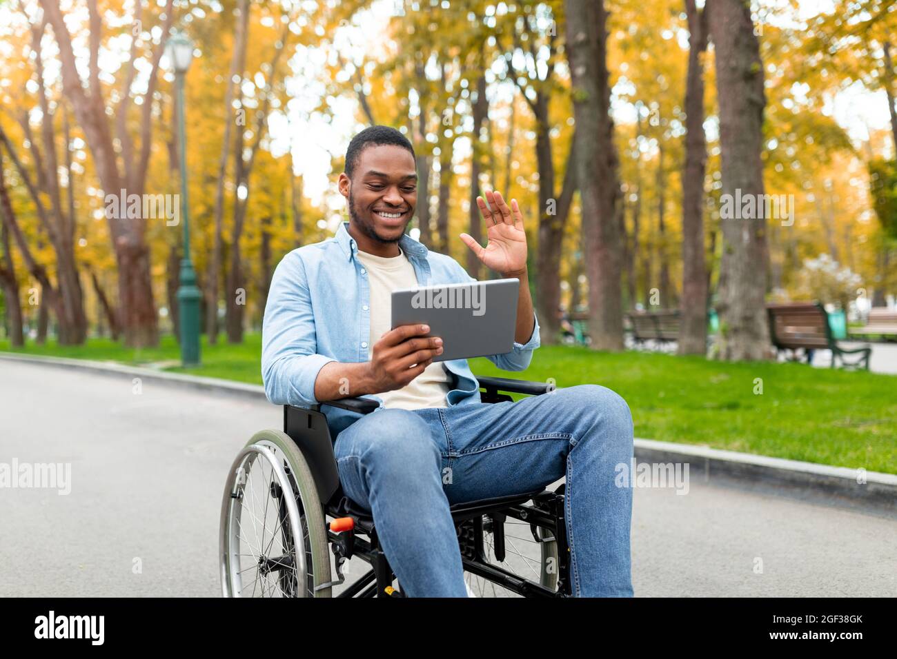 Behinderter schwarzer Mann im Rollstuhl, der Tablet-Computer für die Fernkommunikation im Herbstpark benutzte und vor der Webcam winkte Stockfoto