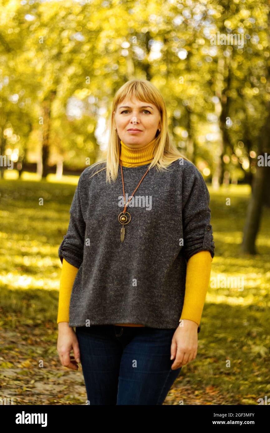 Defokussieren Licht lächelnde blonde 40er Frau im gelben Herbst Park stehen. Frohe, schöne Dame. Frauen tragen grauen Pullover, gelben Rollkragen, Anhänger Stockfoto