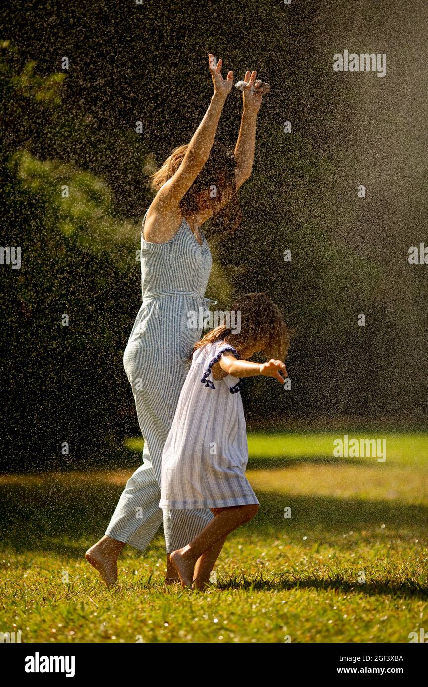 Nettes kleines Mädchen, das mit ihrer Mutter Spaß mit Wasser unter Bewässerungsregner hat Stockfoto