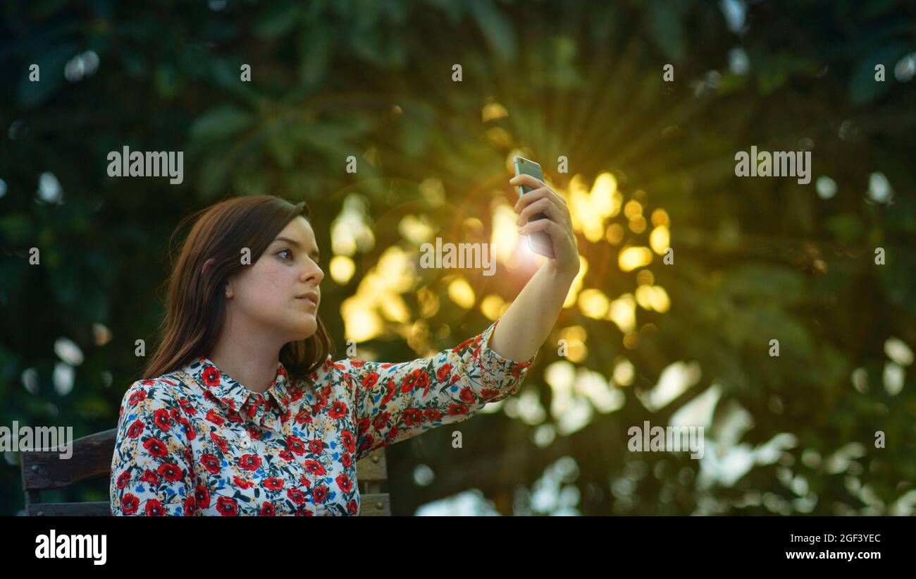Schöne junge lateinerin im Kleid mit Blumenmuster, die bei Sonnenuntergang mit ihrem Smartphone ein Selfie im Garten macht, wobei die Sonnenstrahlen durch den ziehen Stockfoto