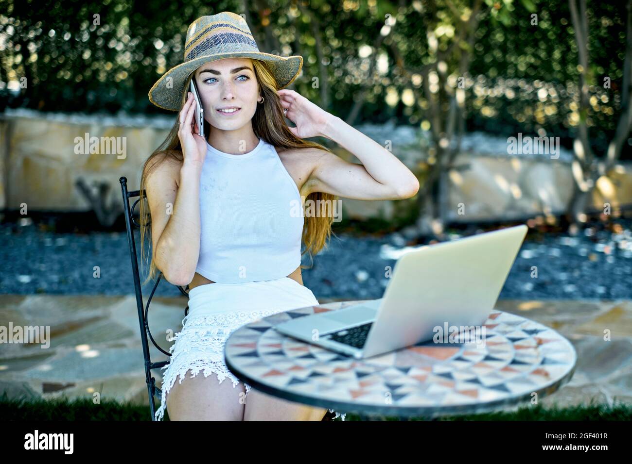 Porträt einer jungen, schönen kaukasischen Frau in den Zwanzigern, die einen Hut trägt und im Freien in einem Garten mit ihrem Mobiltelefon und einem Laptop auf der Suche ist Stockfoto