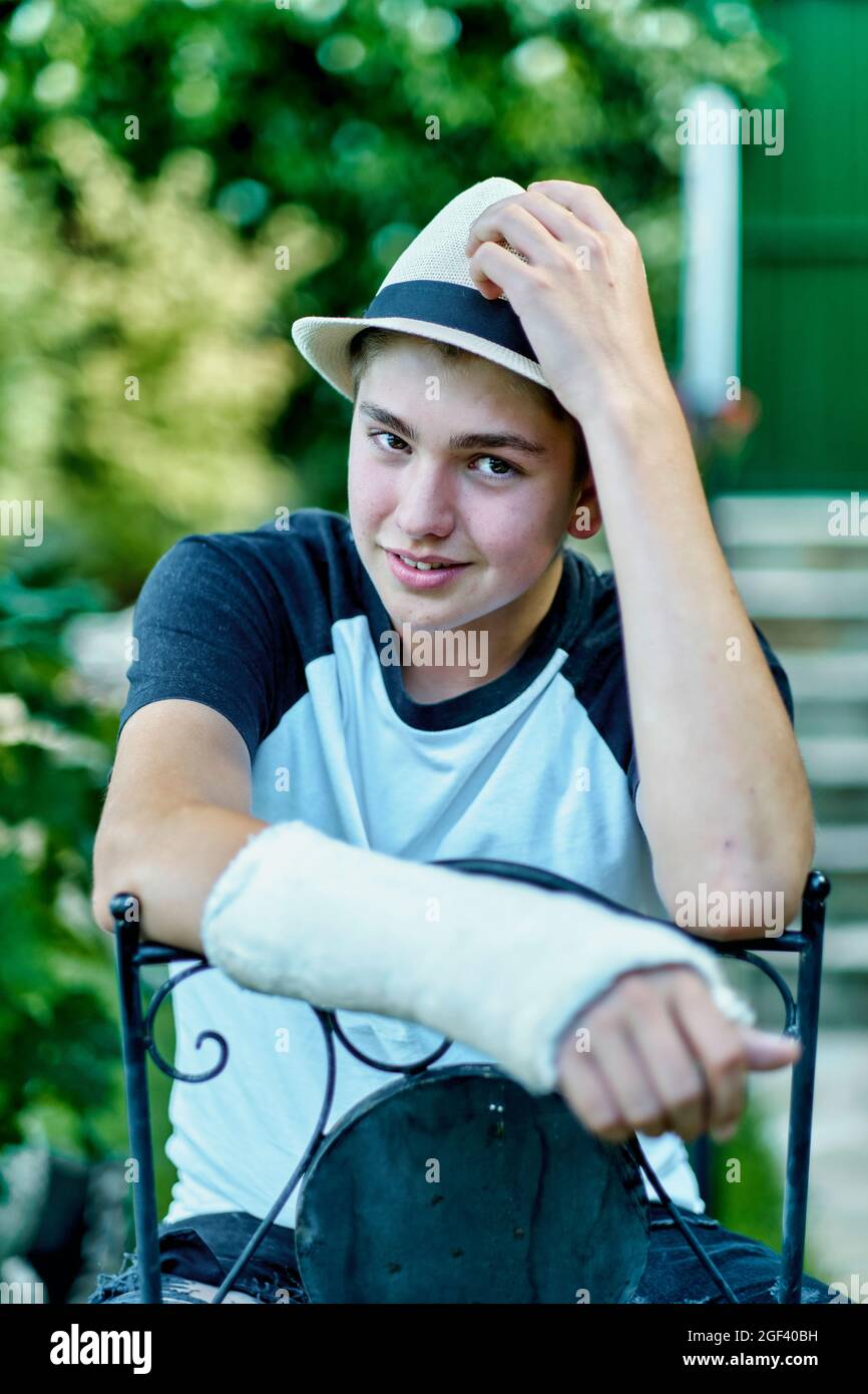 Porträt eines jungen kaukasischen Jungen mit einem gebrochenen und gegossenen Arm, der einen Hut trägt und auf einem Stuhl im Garten sitzt. Lifestyle-Konzept. Stockfoto