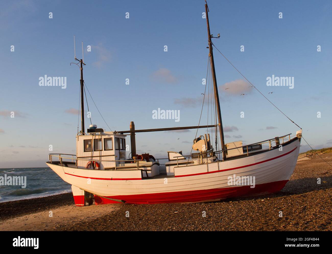 Rot-weißer Küstenkutter im warmen Licht des Sonnenuntergangs, gezeichnet am Strand von Lild Strand in Dänemark Stockfoto