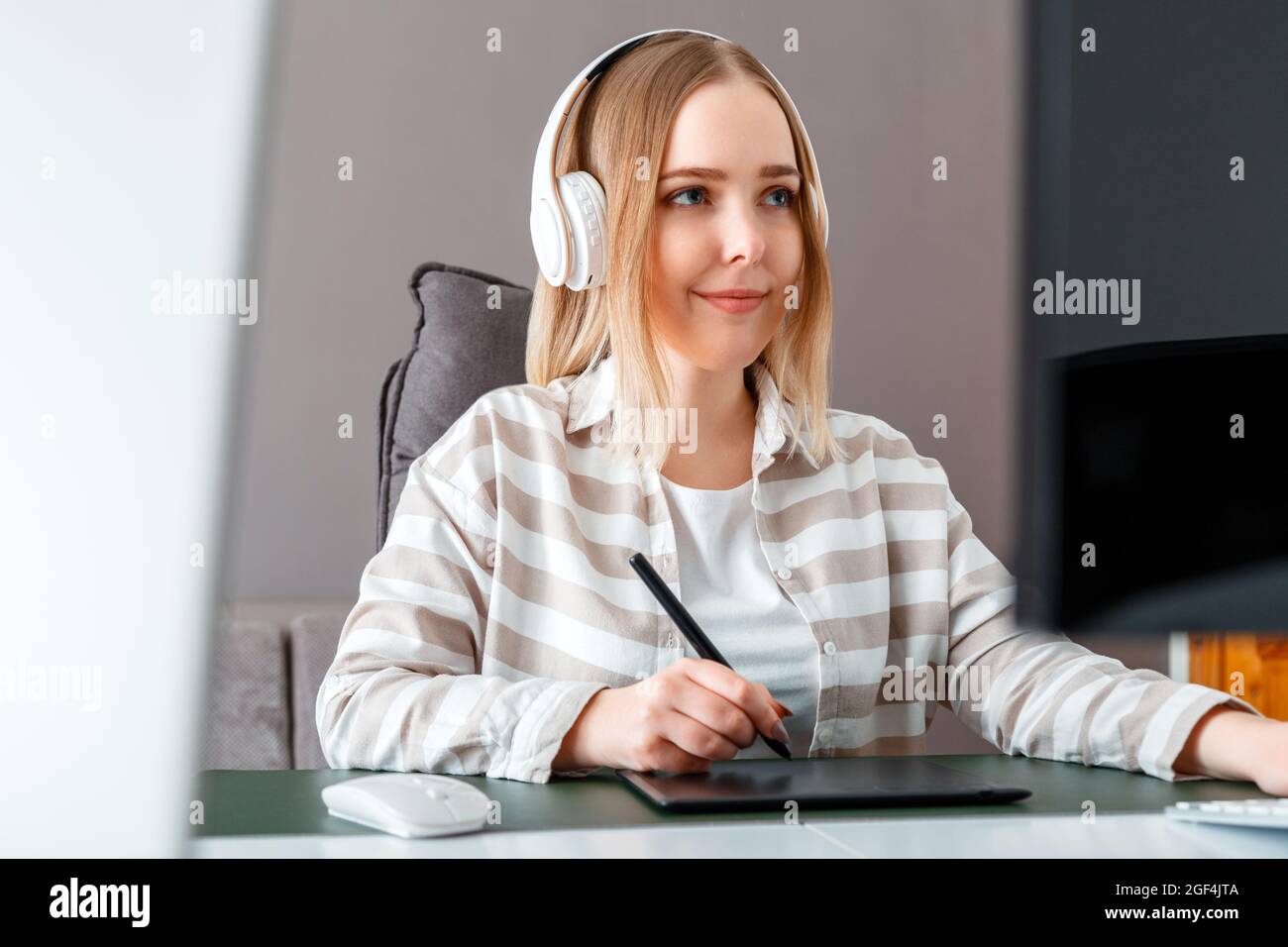 Junge freiberufliche Designerin verwendet während der Arbeit ein Grafiktablett. Digitale Künstlerin Frau in Kopfhörern arbeitet mit pc-Computer im Home Office. Entfernt Stockfoto