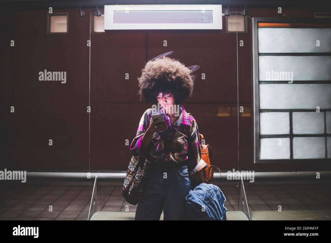 Junge Frau mit Afro-Frisur mit Smartphone an der Bushaltestelle in der Stadt Stockfoto