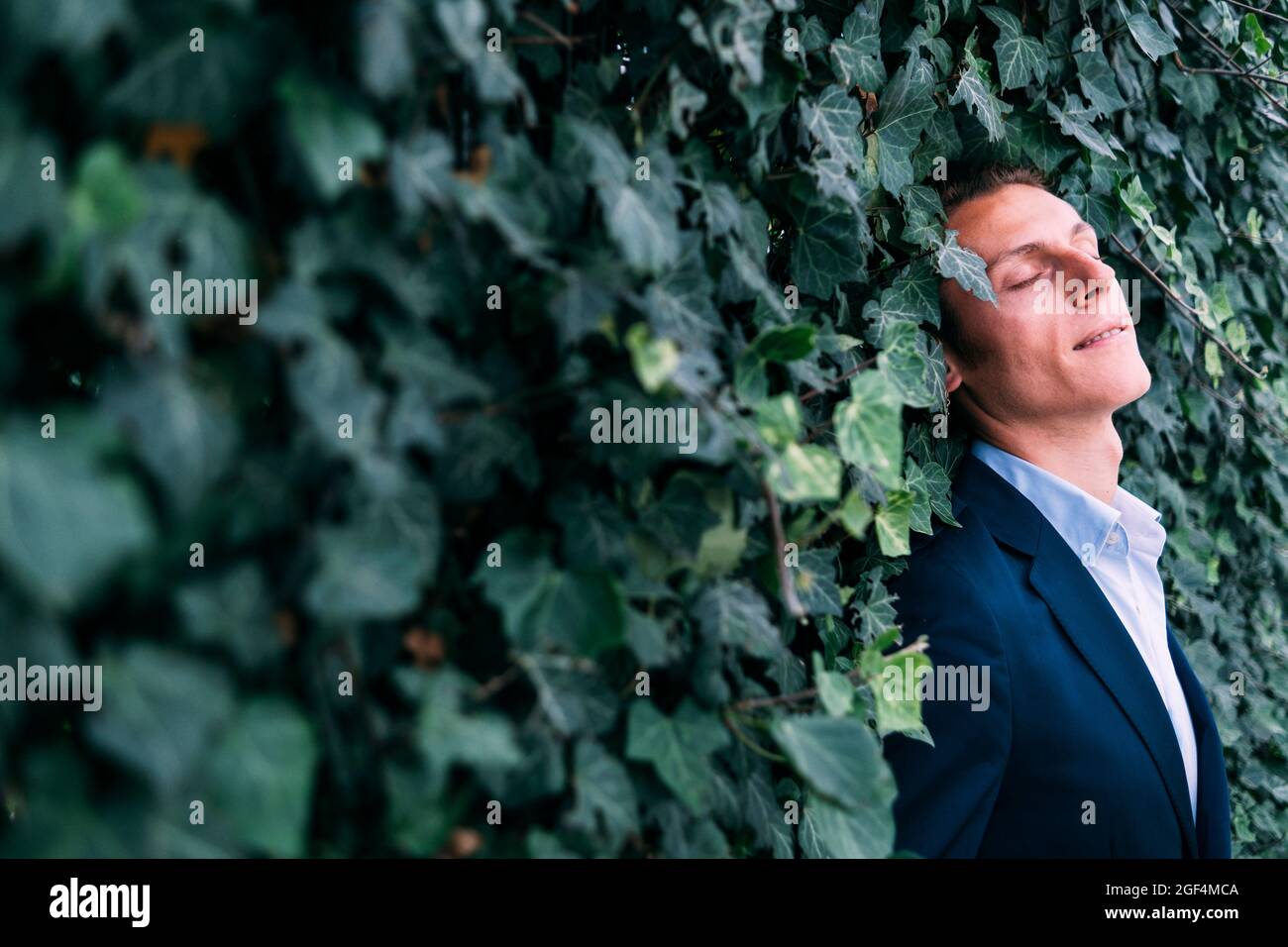 Lächelnder Geschäftsmann, der sich an einer grünen Pflanzenwand lehnt Stockfoto