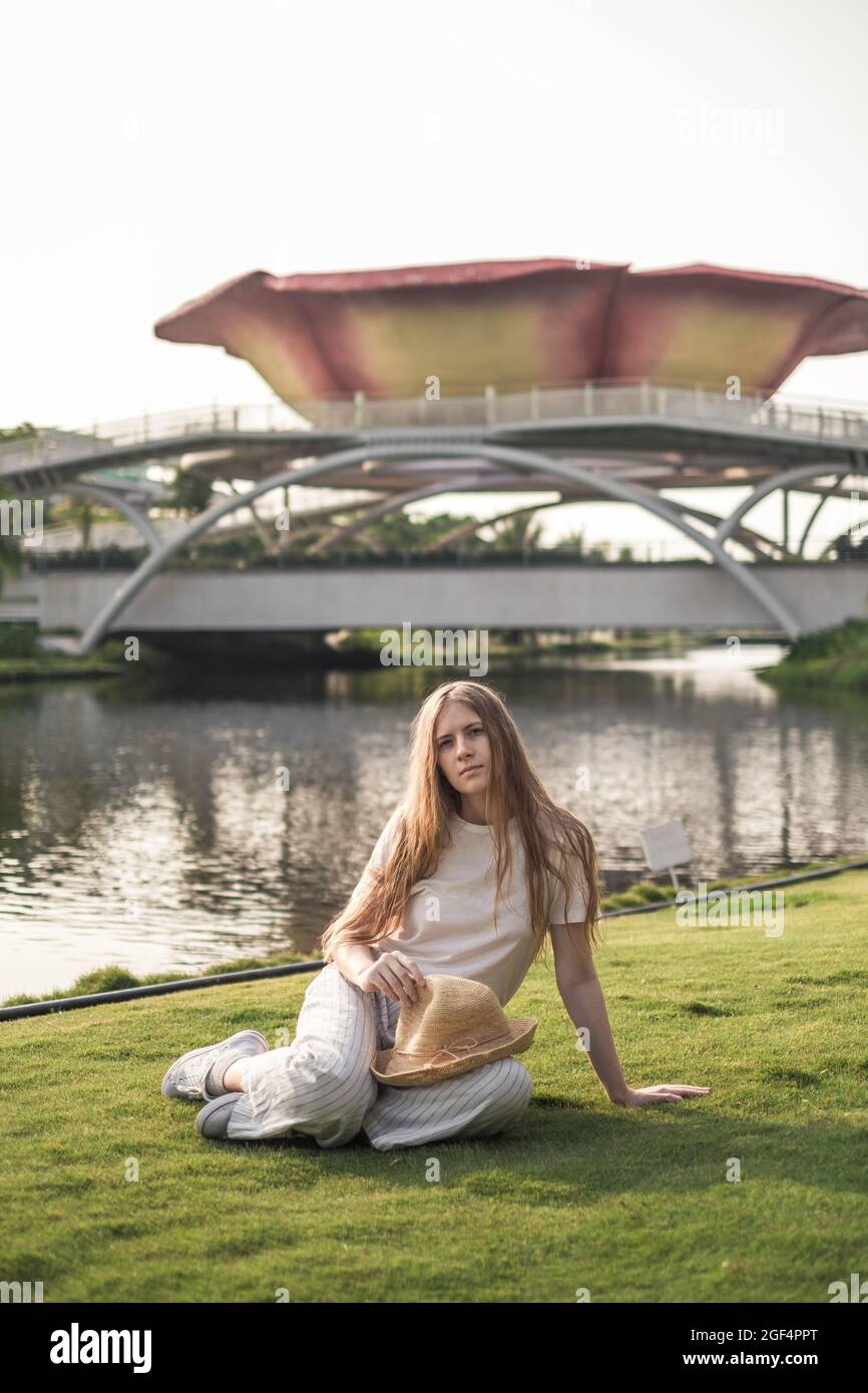 Junge schöne blonde Frau in weißen Freizeitkleidung sitzt auf dem Gras und Blick auf die Kamera mit Kopieplatz. Modisches, lässiges Sommer- oder Frühlingsoutfit Stockfoto