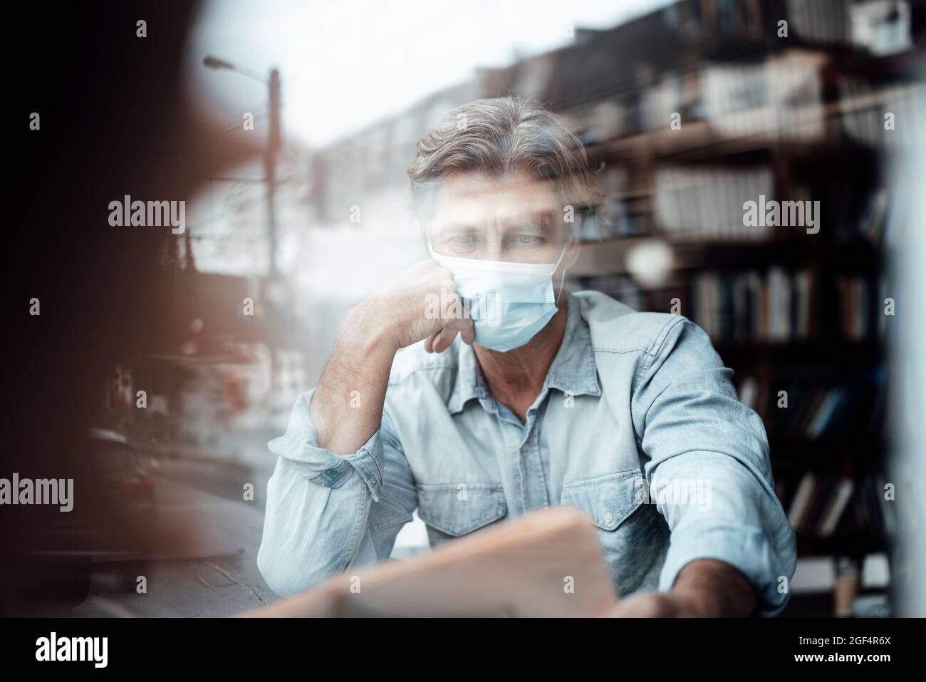 Reifer Mann, der eine schützende Gesichtsmaske trägt, liest Zeitung im Café, das durch das Glasfenster gesehen wird Stockfoto