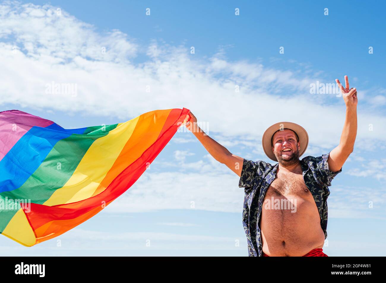Fröhlicher schwuler Mann, der an sonnigen Tagen die Regenbogenfahne unter dem Himmel schwenkt Stockfoto