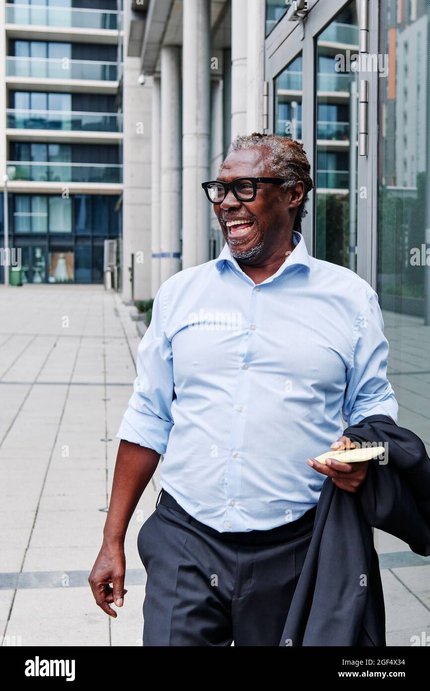 Fröhlicher älterer männlicher Profi mit Handy und Blazer, der auf dem Fußweg läuft Stockfoto