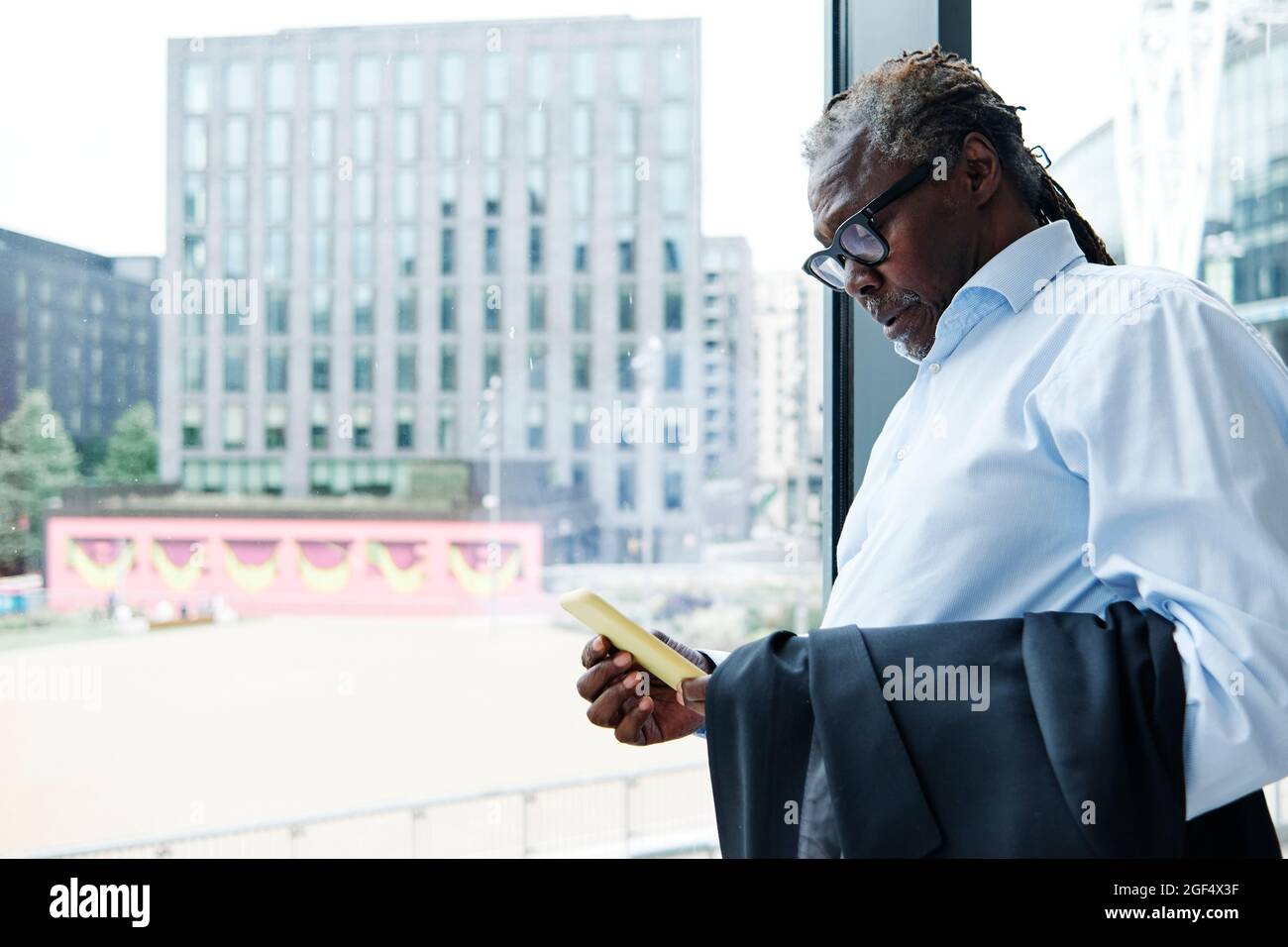 Leitender männlicher Profi, der Smartphone benutzt, während er sich an einer Glaswand lehnt Stockfoto