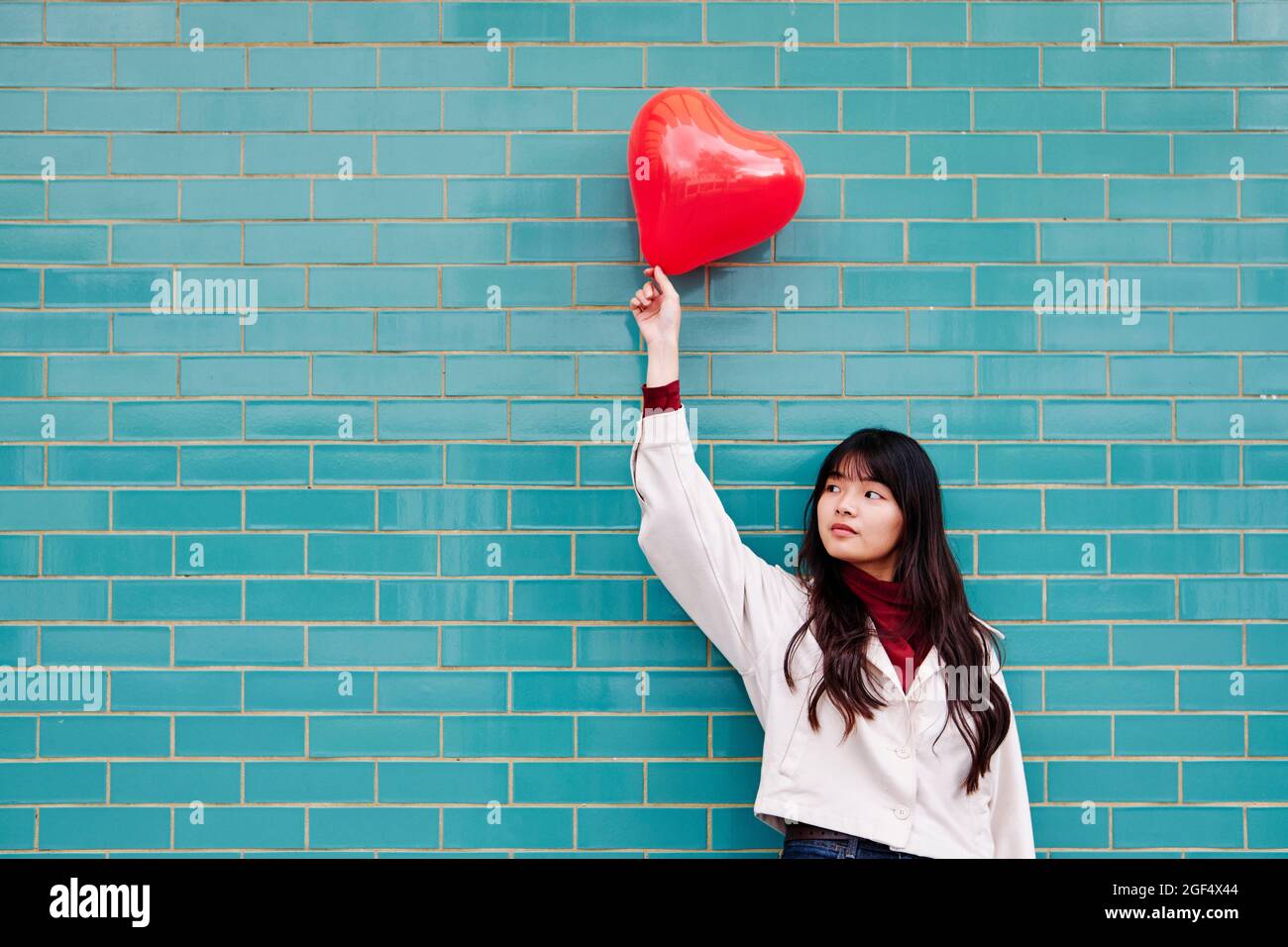 Junge Frau schaut weg, während sie den roten Herzballon vor der Ziegelwand hält Stockfoto