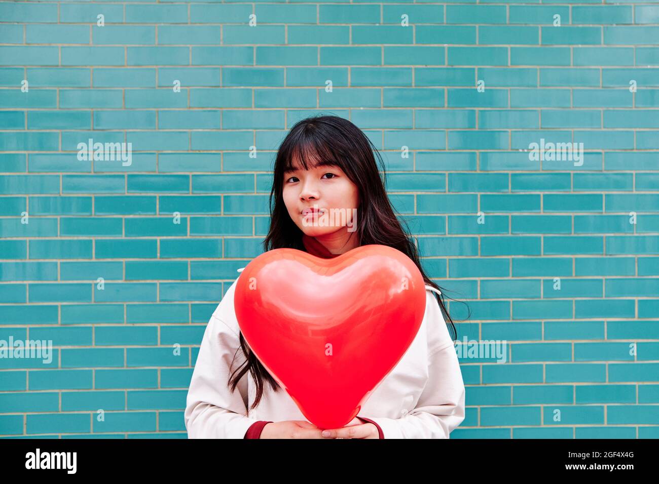 Junge Frau hält einen roten Herzballon in der Nähe einer Ziegelwand Stockfoto