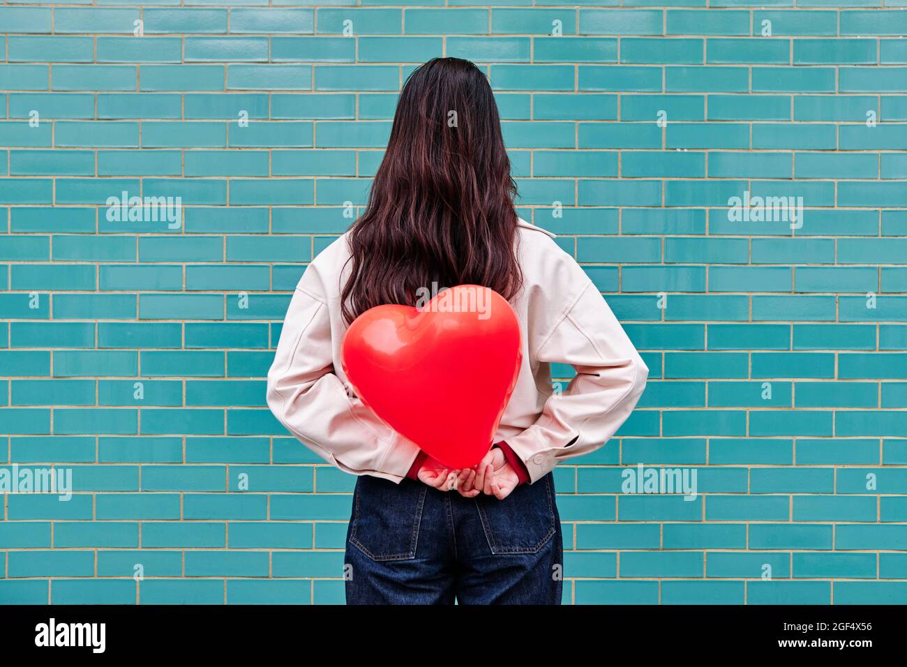 Junge Frau hält einen roten Herzballon hinter ihrem Rücken vor einer Ziegelwand Stockfoto