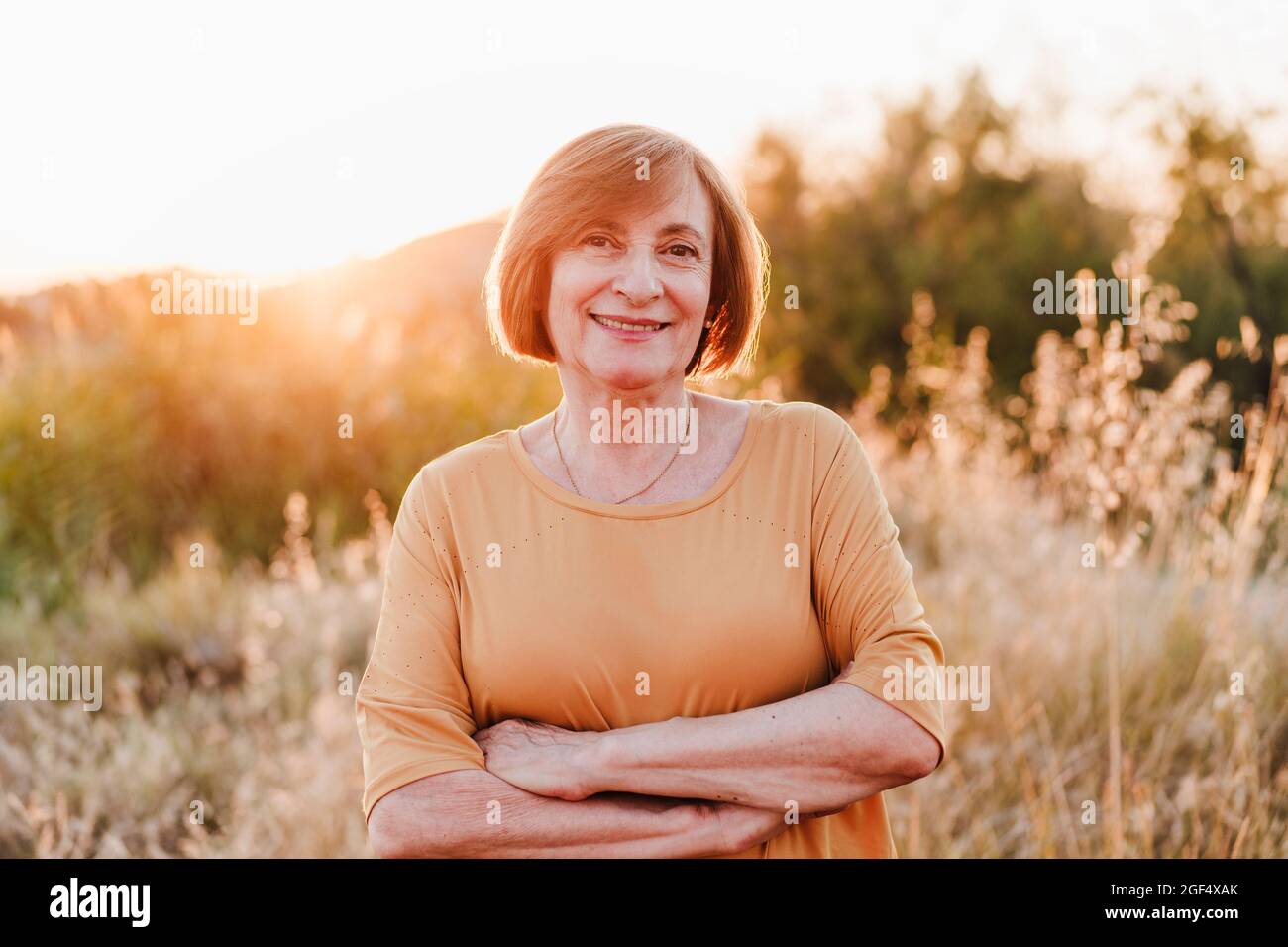 Lächelnde ältere Frau mit gekreuzten Armen während des Sonnenuntergangs Stockfoto