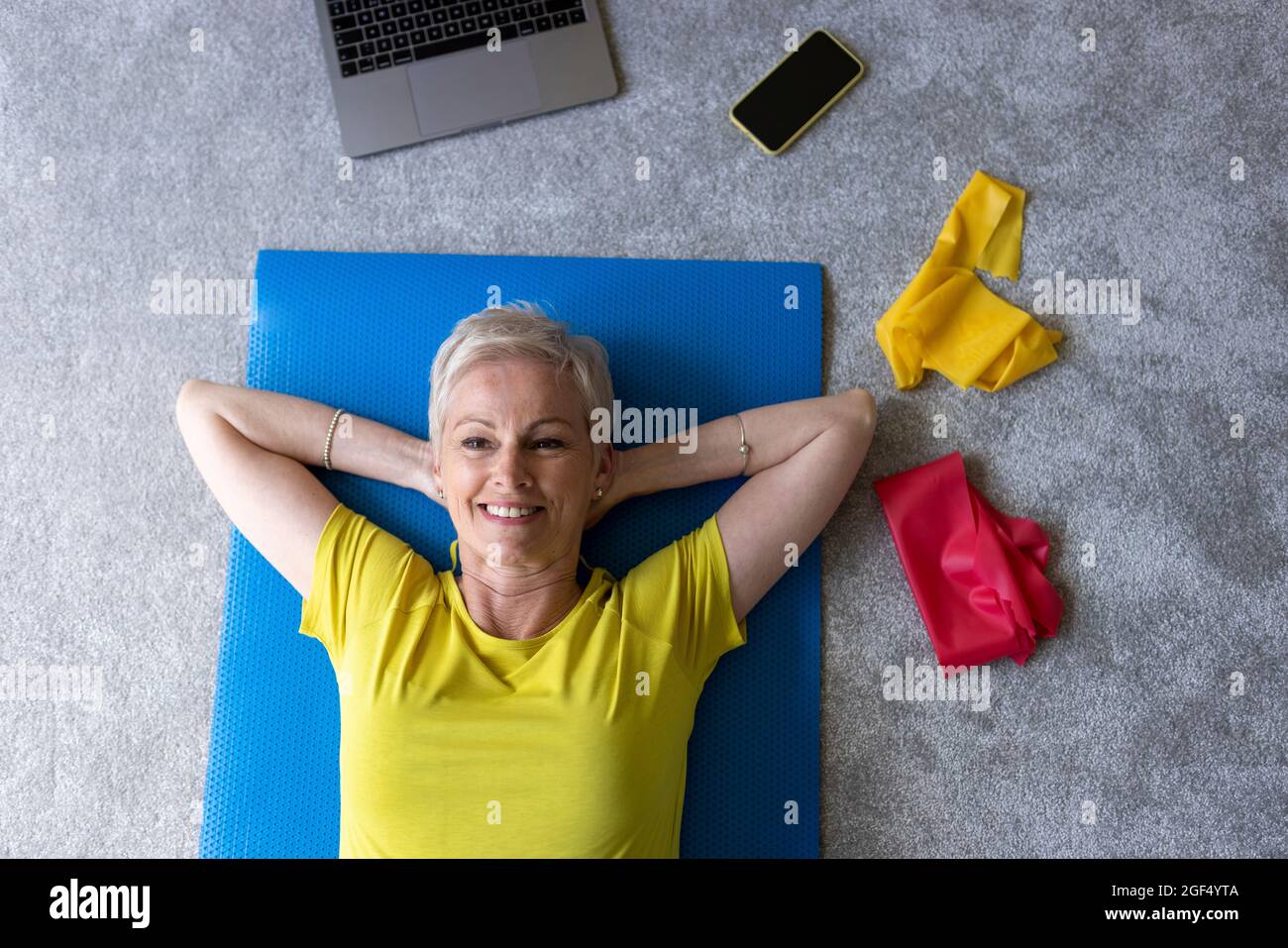 Lächelnde reife Frau mit Händen hinter dem Kopf, die sich auf der Trainingsmatte entspannt Stockfoto