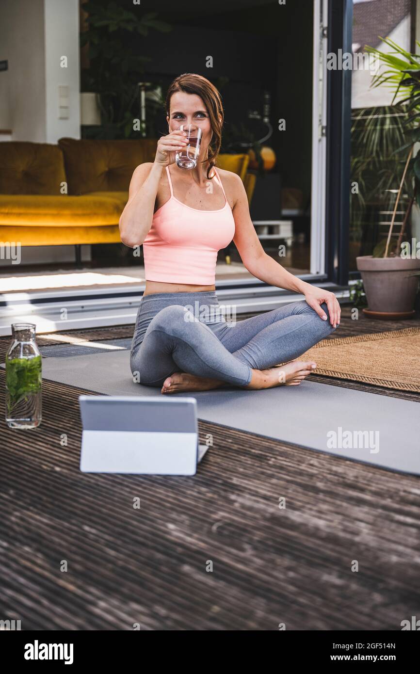 Frau trinkt Entgiftungswasser, während sie auf der Terrasse mit gekreuzten Beinen sitzt Stockfoto