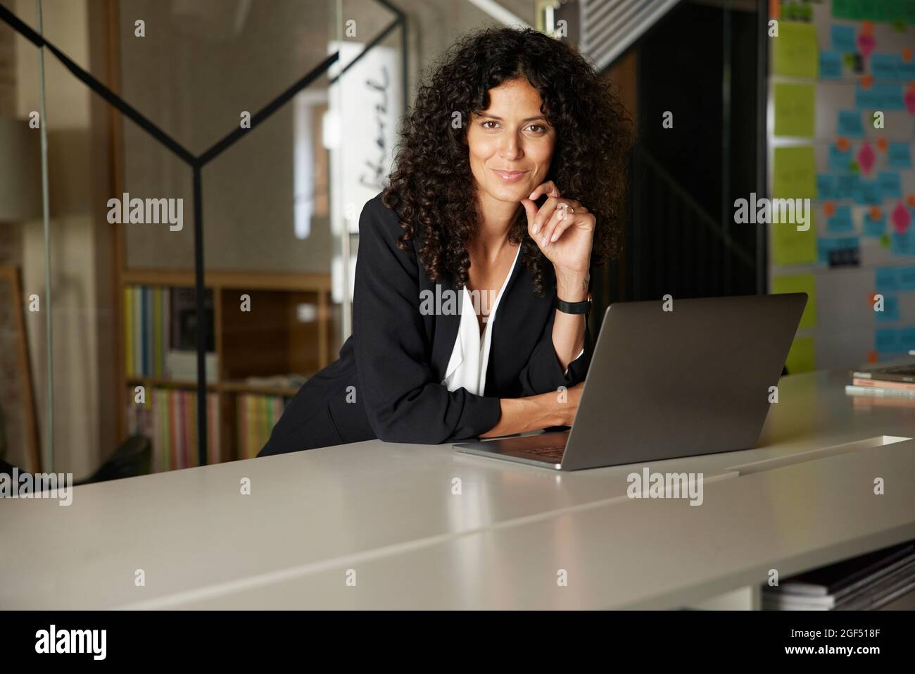 Lächelnde Geschäftsfrau mit der Hand auf dem Kinn am Laptop am Arbeitsplatz Stockfoto