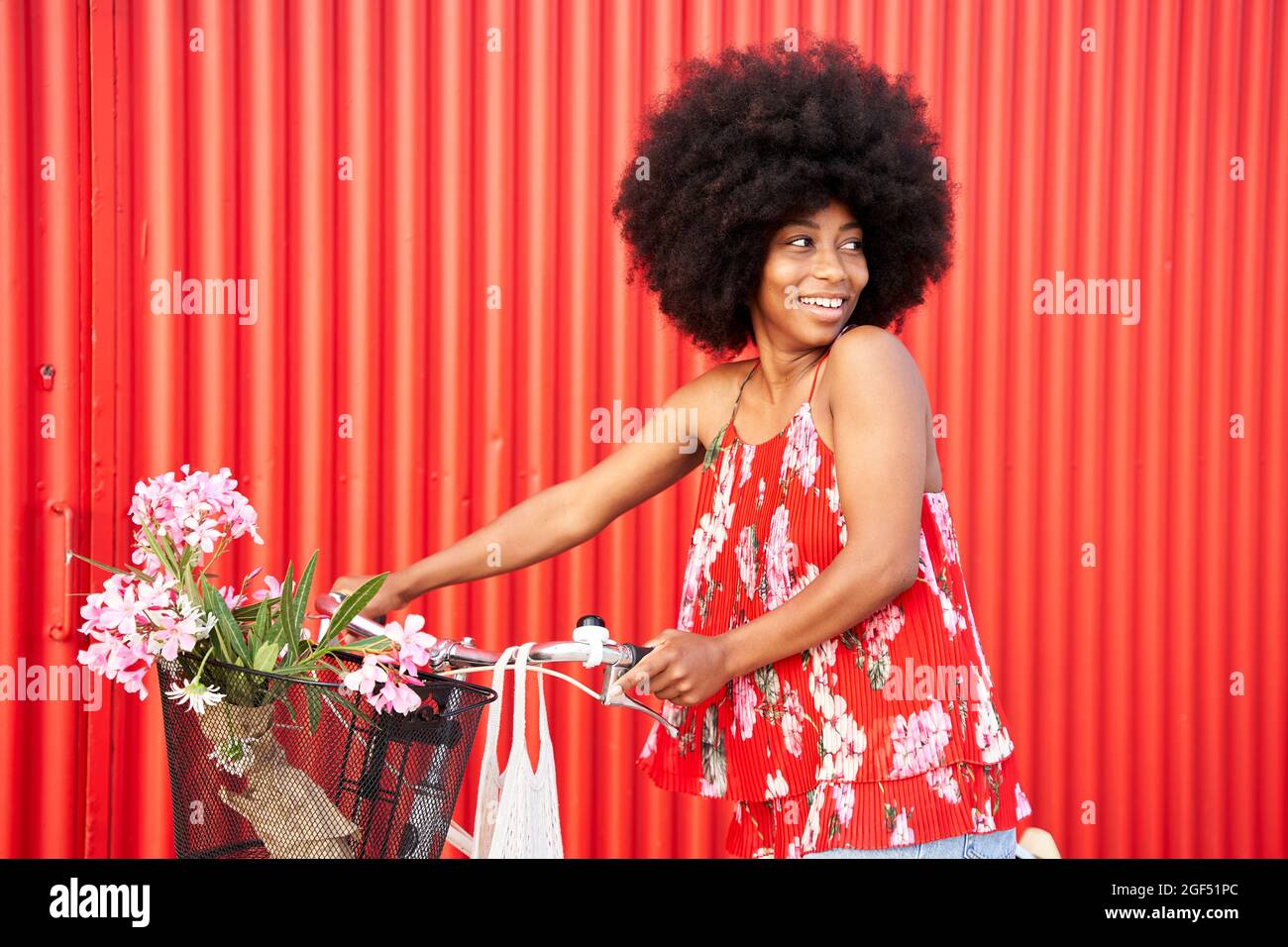 Afro-Frau mit Fahrrad lächelt, während sie an der roten Wand steht Stockfoto